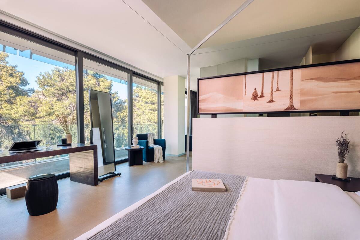 Quarto de hotel moderno com uma janela grande, uma cama, uma escrivaninha com um laptop e uma área de estar com uma cadeira e uma mesinha.