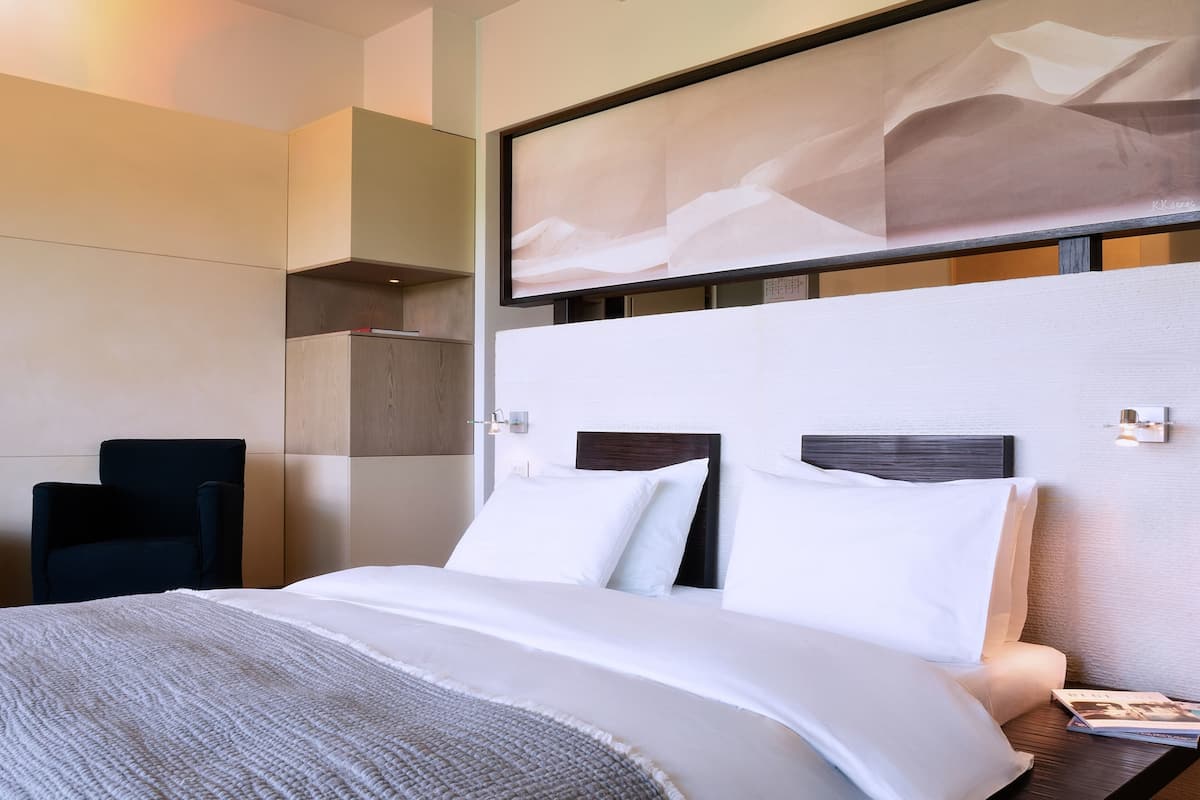 A hotel room with a bed, a chair, a wall-mounted artwork, and a nightstand with a lamp.