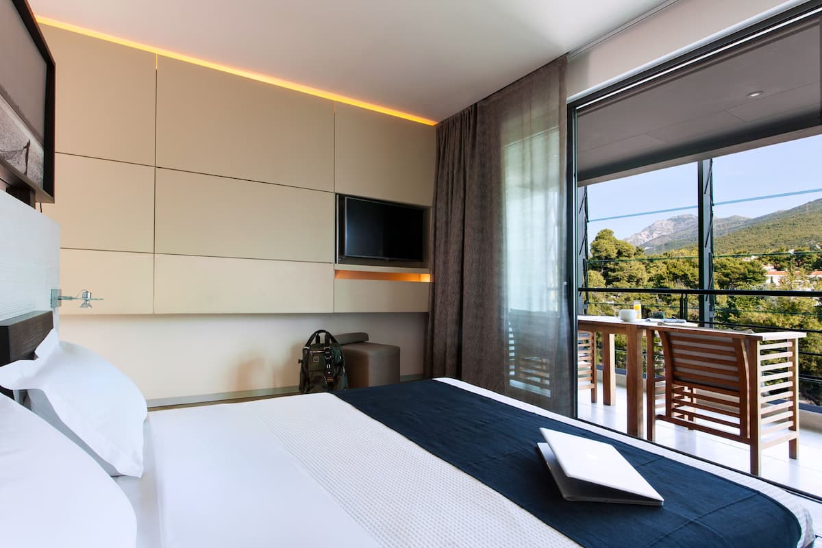 Quarto de hotel moderno com uma cama grande, televisão de tela plana na parede, uma varanda com mesa e cadeiras, e vista panorâmica para as montanhas.