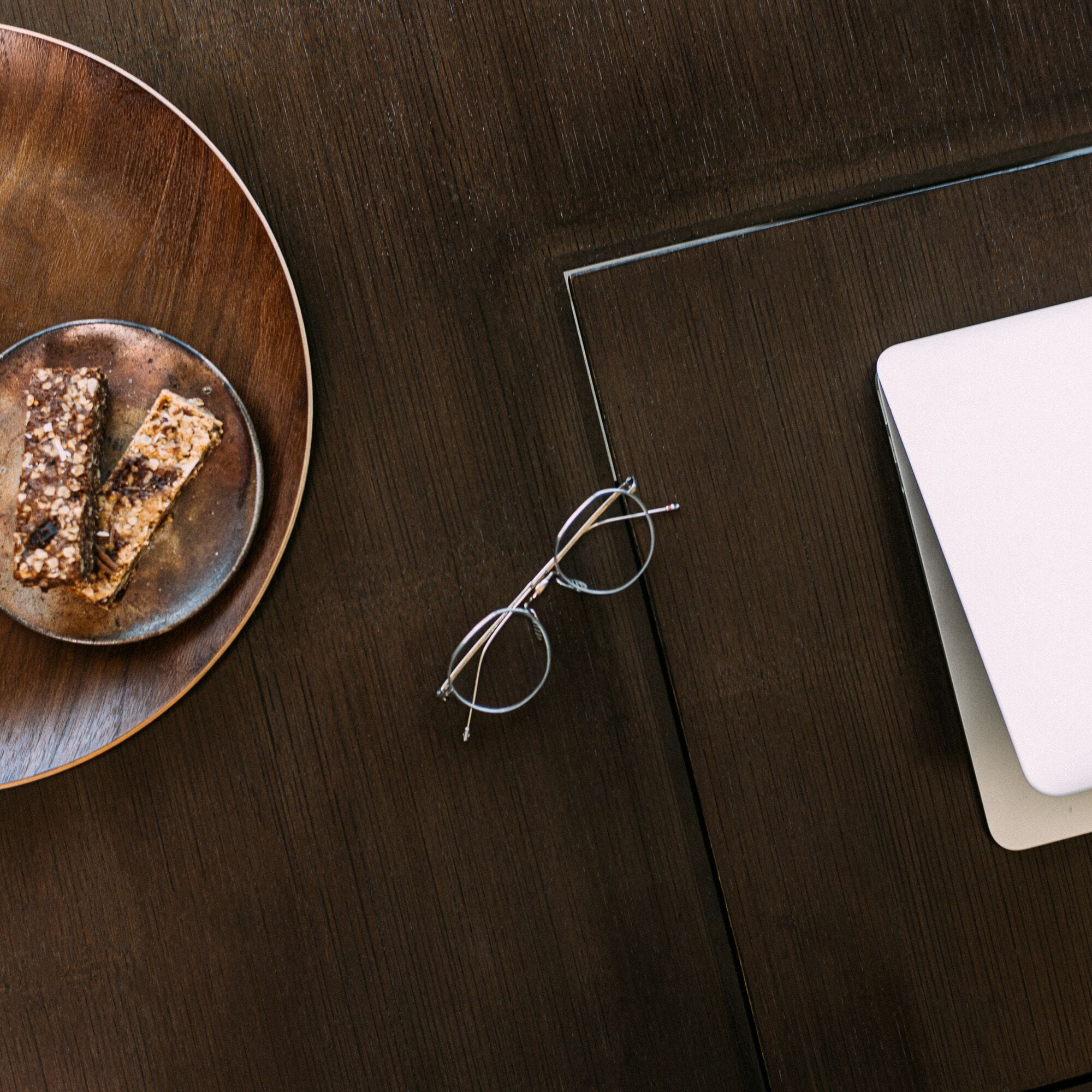 A wooden table with a plate of granola bars, a pair of glasses, and a laptop.