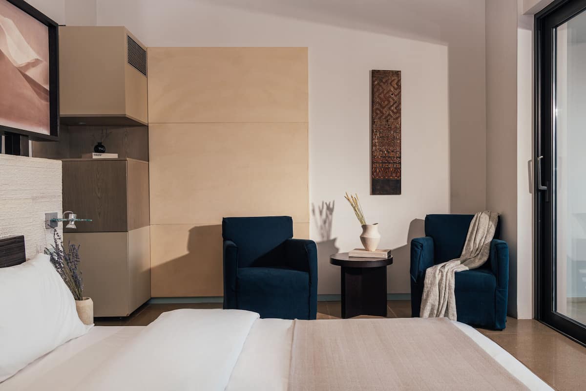 Quarto de hotel moderno com uma cama grande, duas poltronas, uma mesinha de centro com um vaso e um objeto decorativo na parede.
