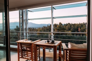 A balcony with a wooden table and chairs, a view of a forest and mountains, and a glass railing.