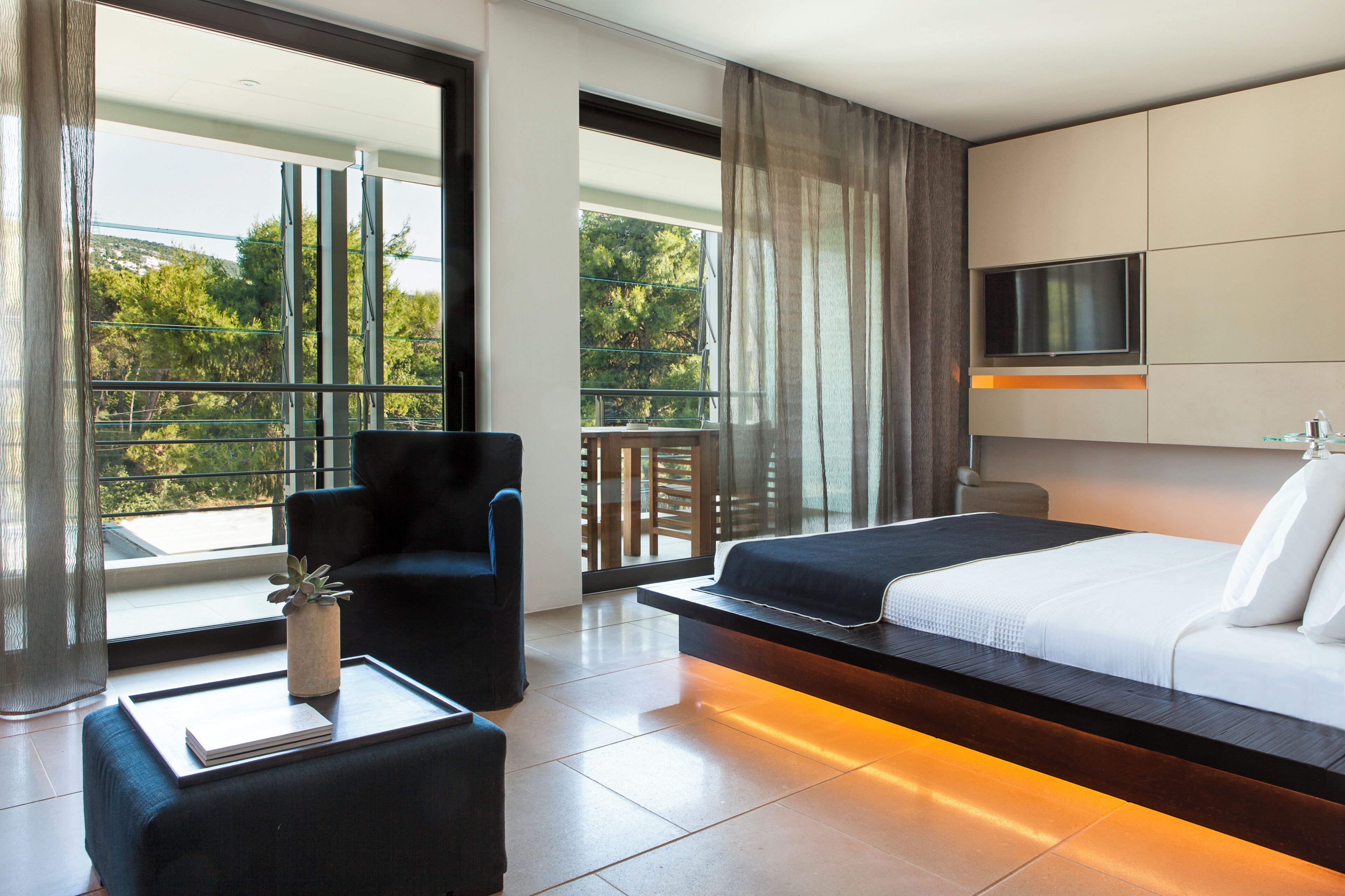 A modern hotel room with a bed, a TV, a chair, a coffee table, and a balcony with a view.