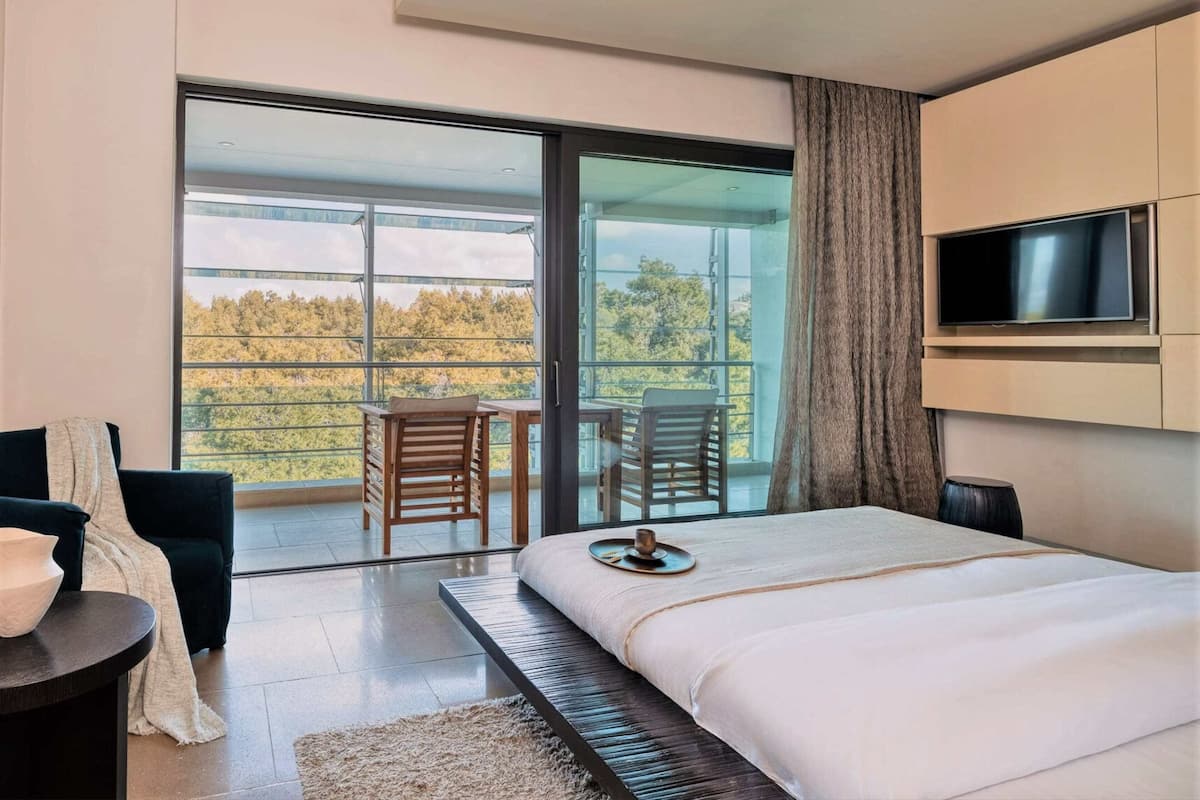 A modern bedroom with a large bed, a TV mounted on the wall, and a balcony with chairs.