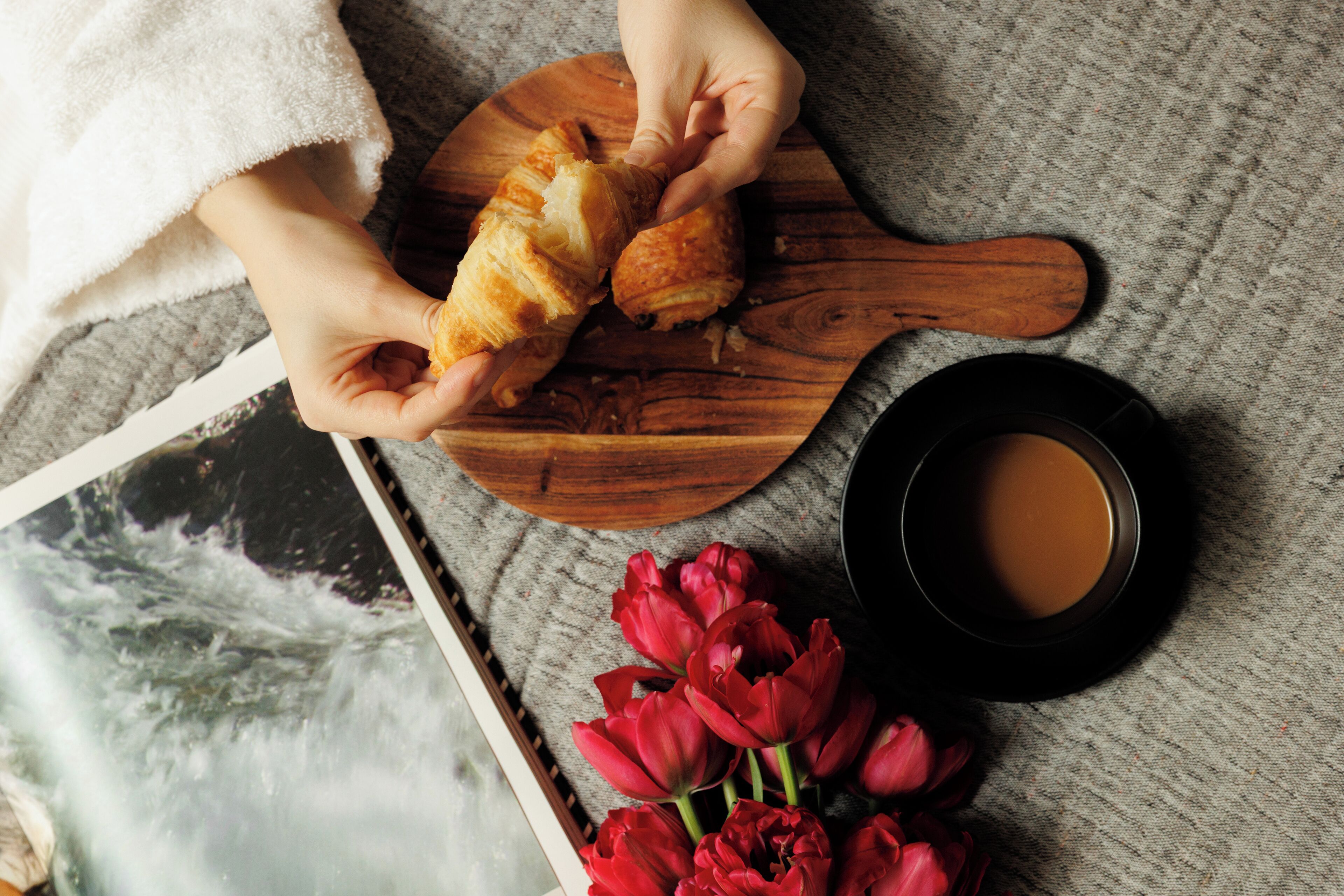 A wooden board with croissants, a black cup, and red tulips on a textured surface.