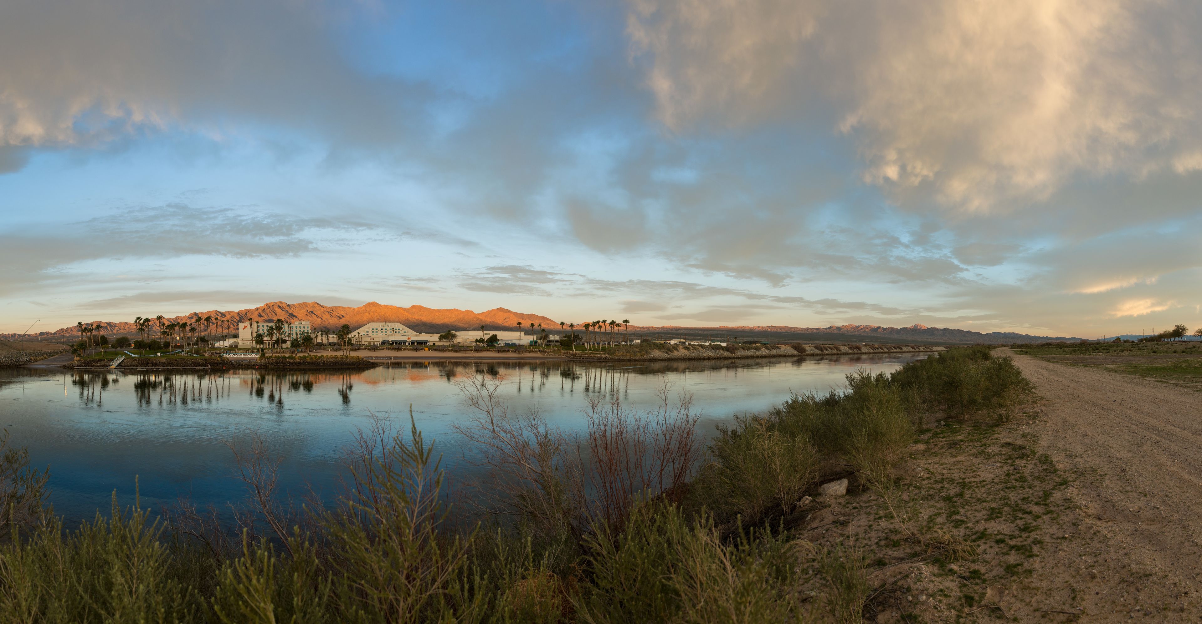 A river with a dirt path alongside, mountains in the background, and a building with a flat roof near the water.