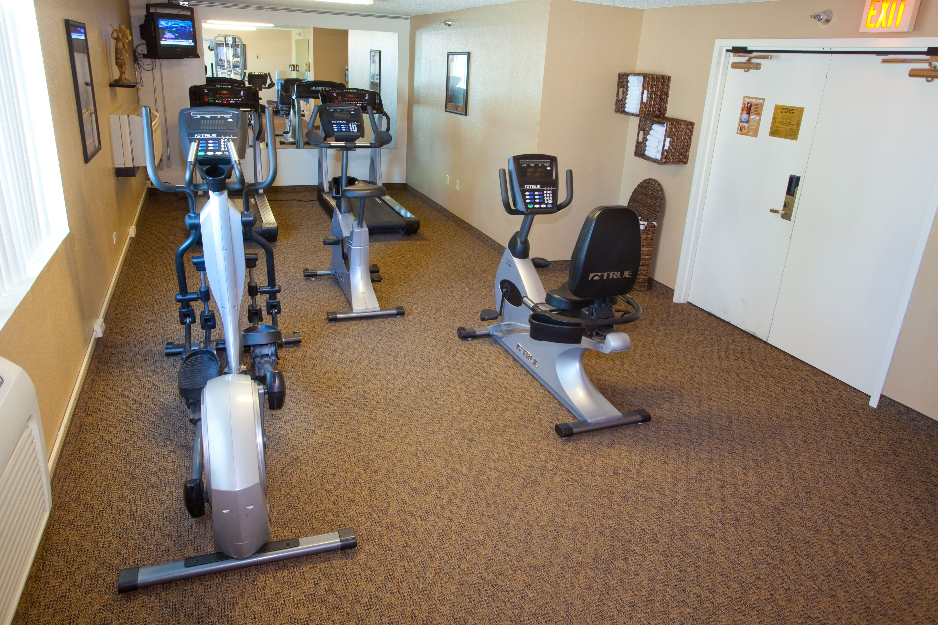 A fitness center with cardio machines, a mirrored wall, and a door with an exit sign.