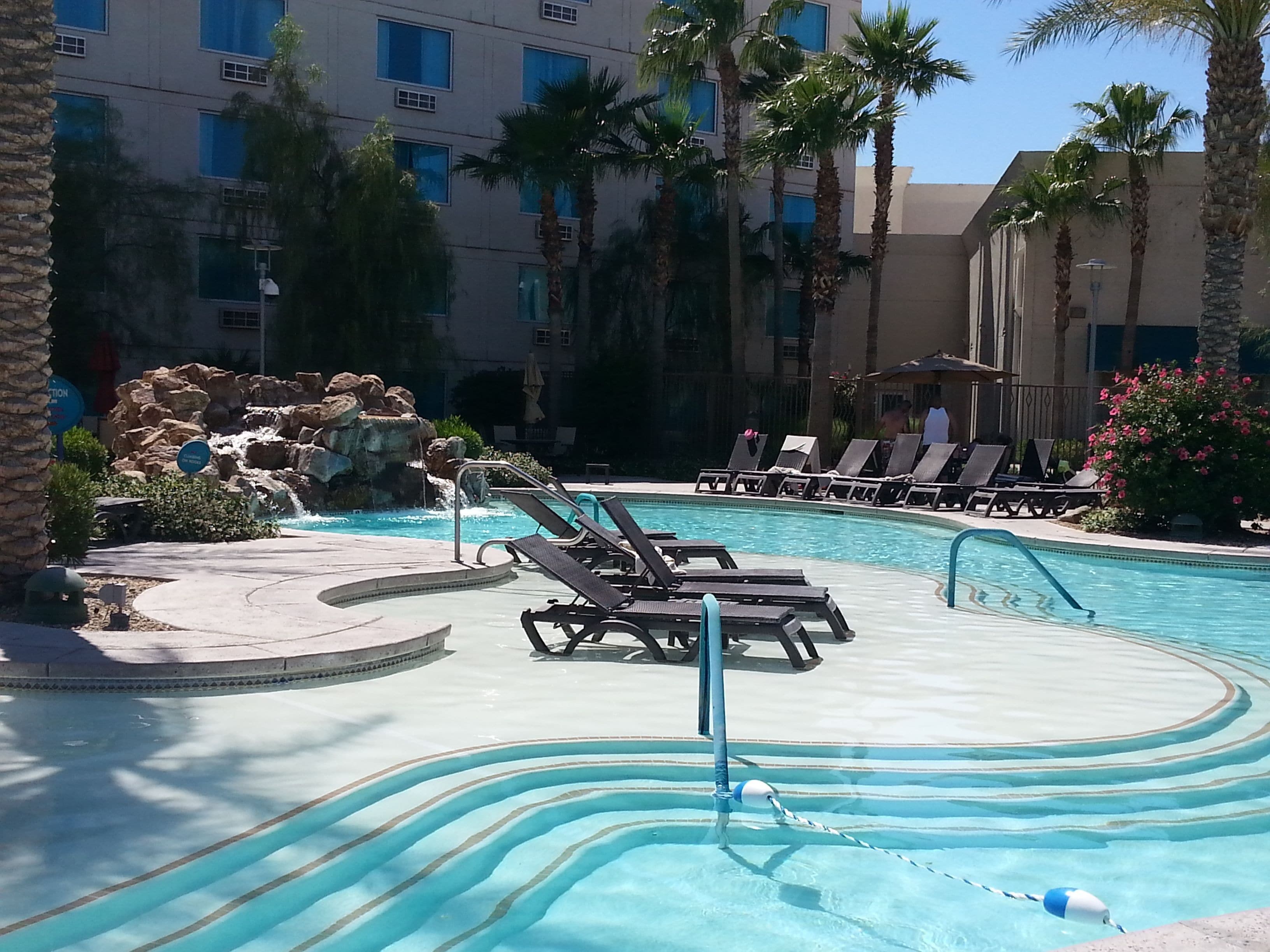 A pool area with lounge chairs, a small waterfall feature, and a building with multiple windows in the background.