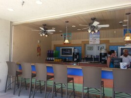 Uma área de bar com várias ventoinhas no teto, um balcão com dispensador de bebidas e uma tela de televisão exibindo um programa.