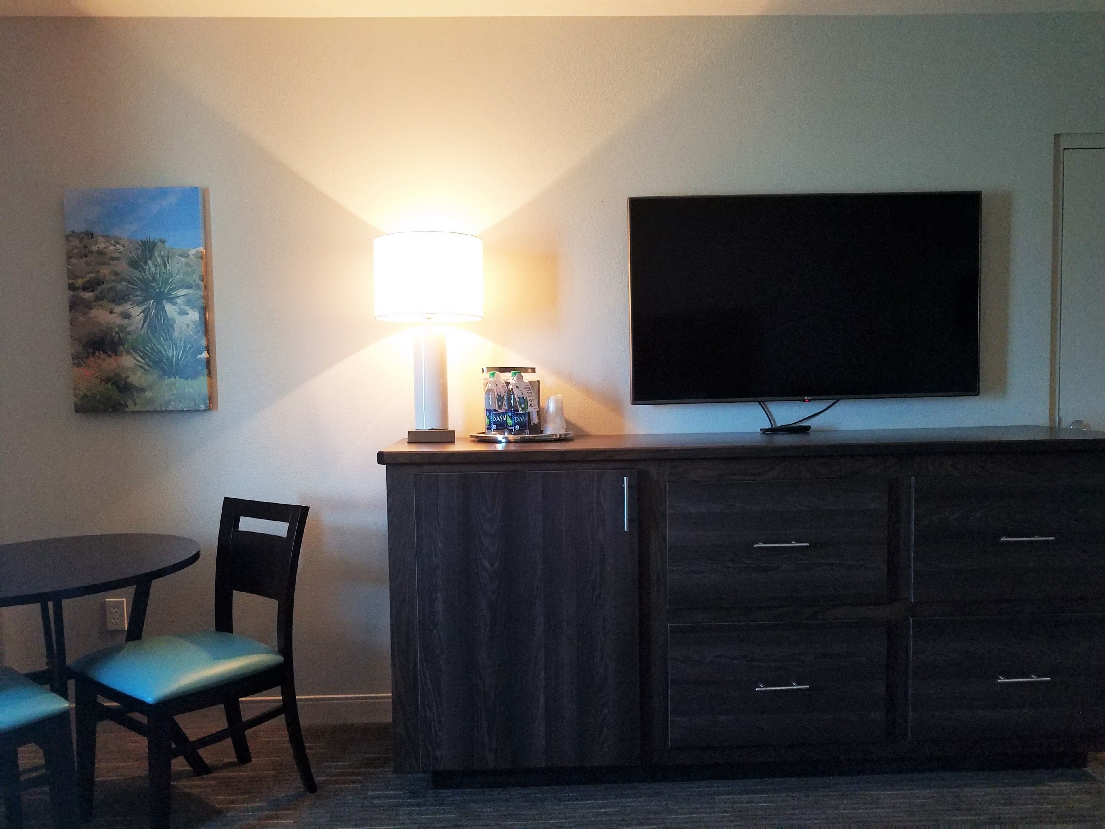 A hotel room with a dark wood cabinet, a flat-screen TV mounted on the wall, a table with two chairs, and a painting on the wall.