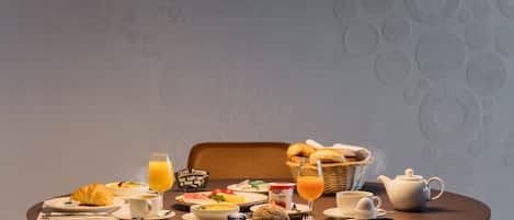 A breakfast setting with a table set for two, including croissants, a basket of bread, a teapot, and a bowl of fruit.