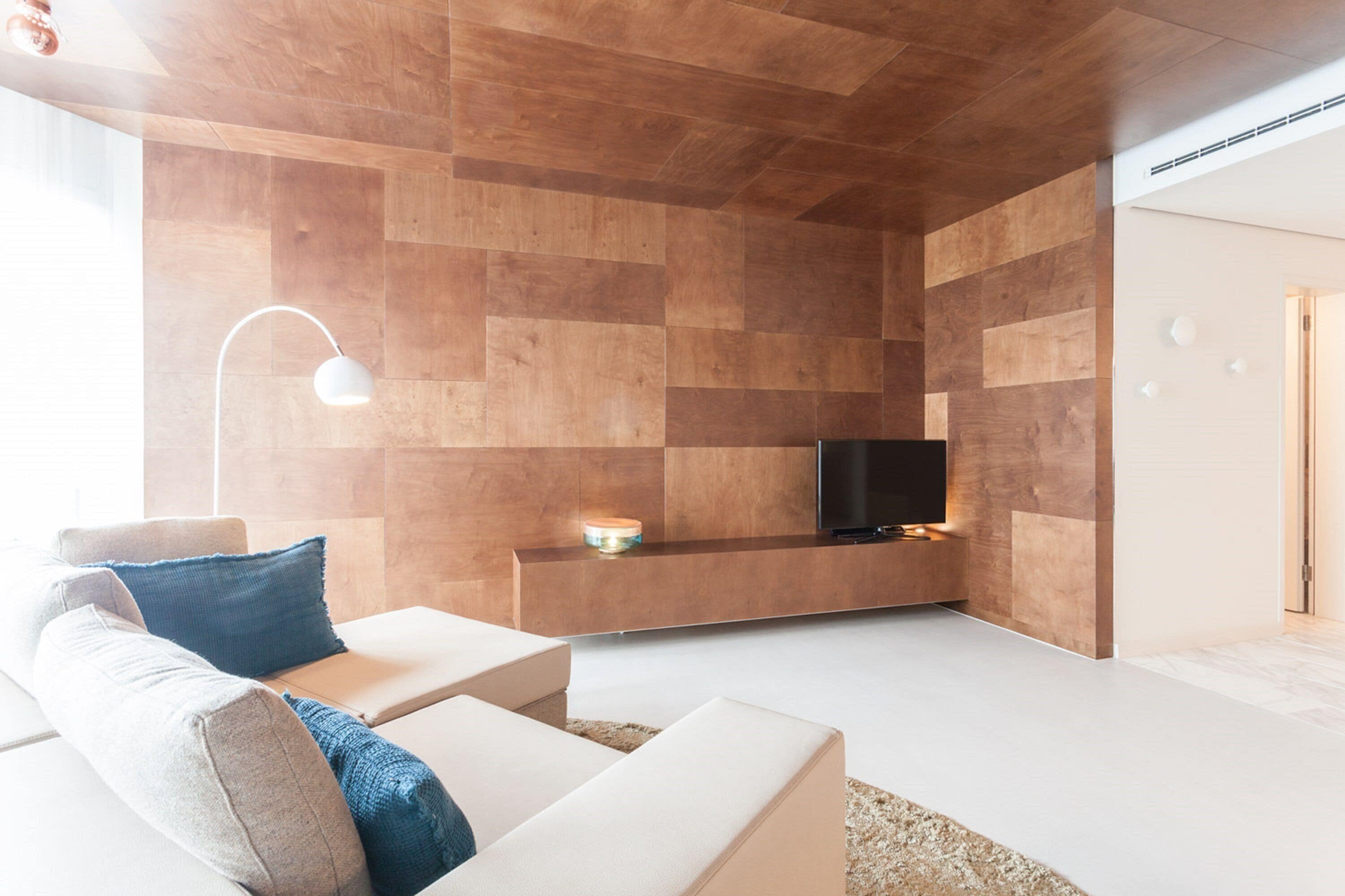 A modern living room with a wooden wall panel design, a flat-screen TV, and a white sofa with blue cushions.