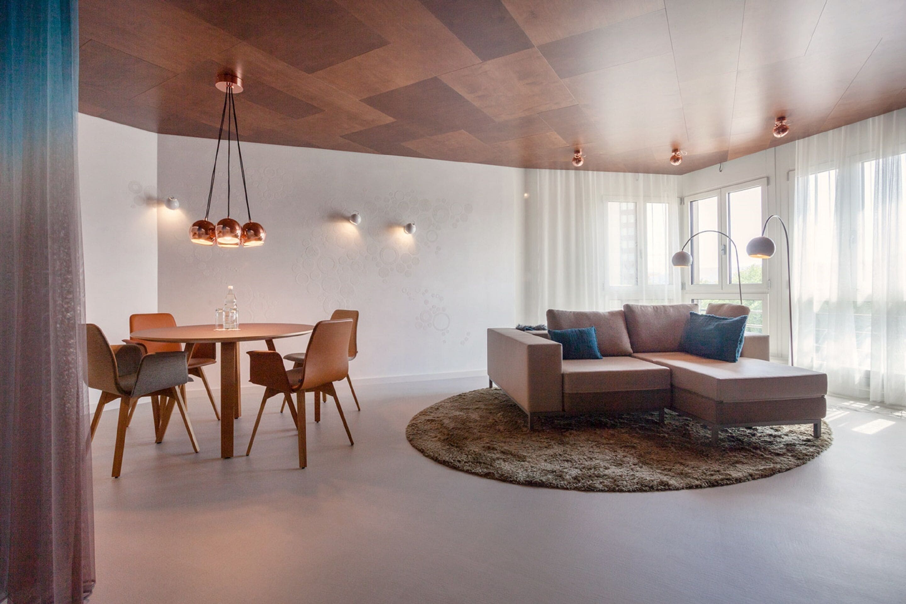 Una sala de estar moderna con una mesa redonda de comedor, un sofá seccional y un ventanal con cortinas de gasa.