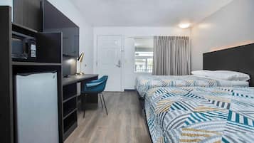 Habitación de hotel compacta con kitchenette, escritorio y silla, dos camas con ropa de cama estampada y vista al exterior a través de una puerta.