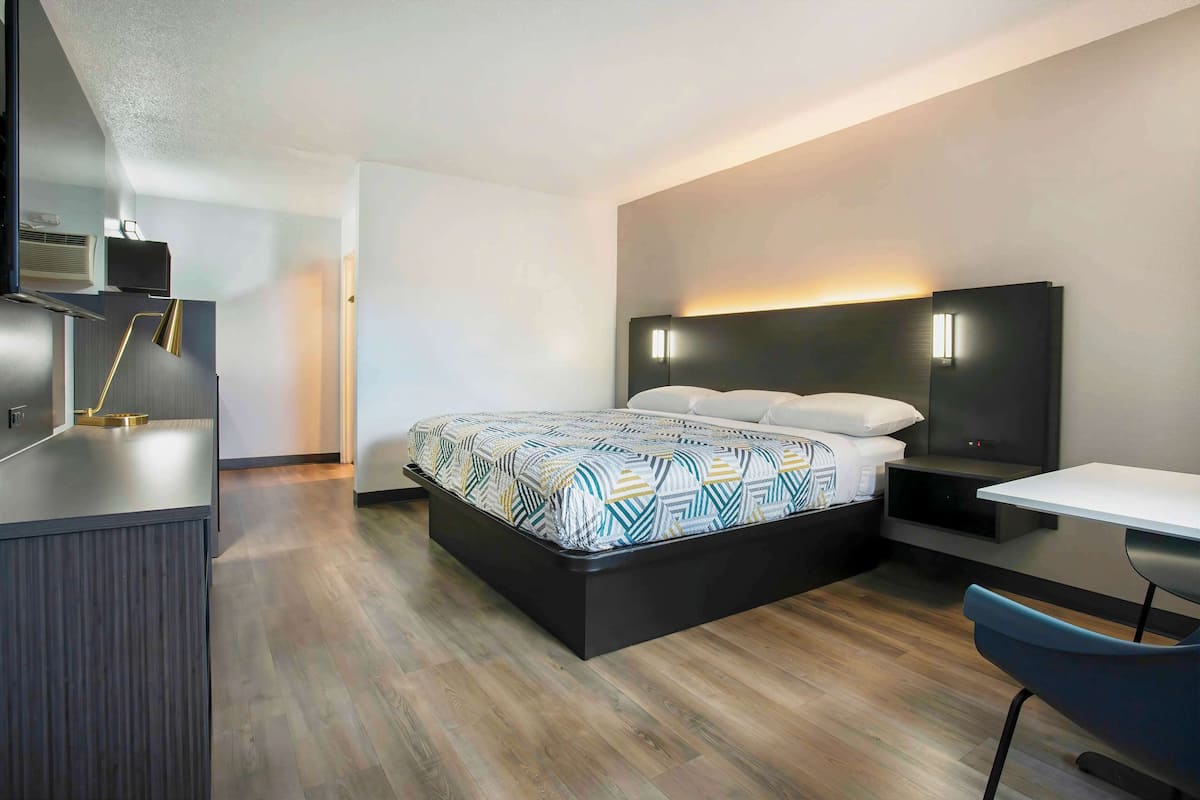 A modern hotel room with a bed, bedside table, desk, and a built-in TV.