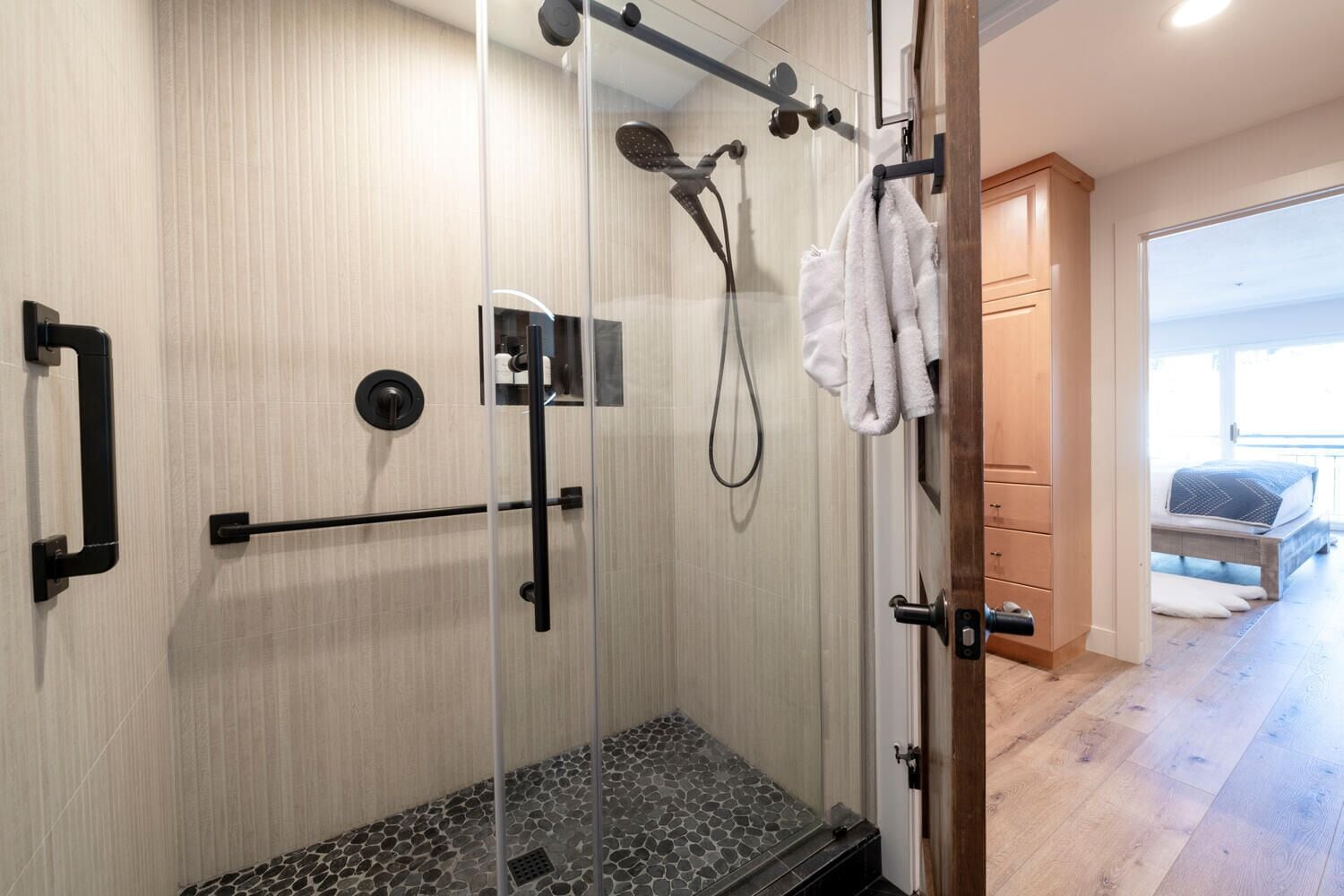 Une salle de bain moderne dotée d’une douche à parois de verre, d’un porte-serviettes et d’une chambre visible avec un lit et une fenêtre.