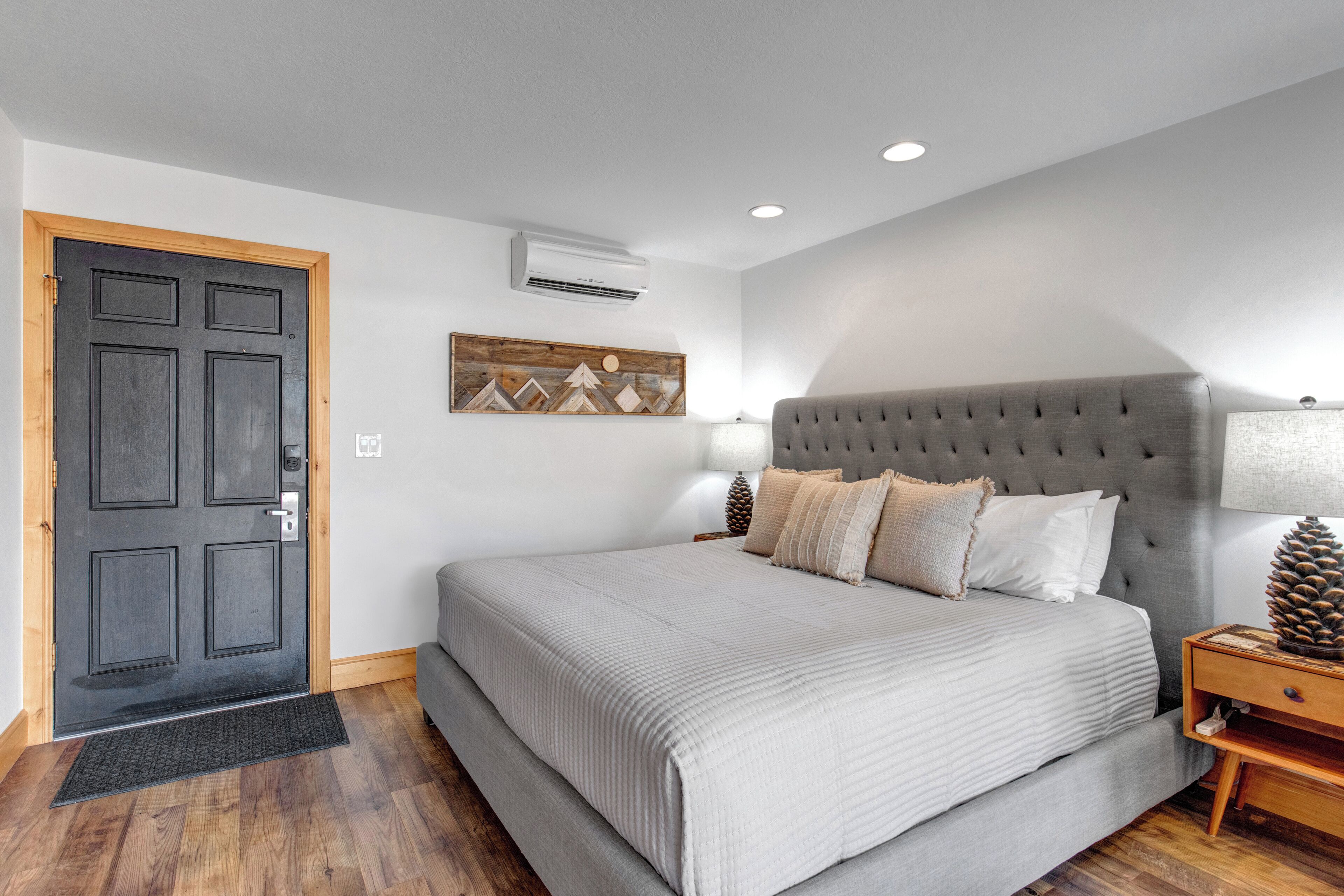 Un dormitorio con una cama grande, una puerta de madera, aire acondicionado, una obra de arte en la pared y una mesita de noche con una lámpara y una decoración de piña.