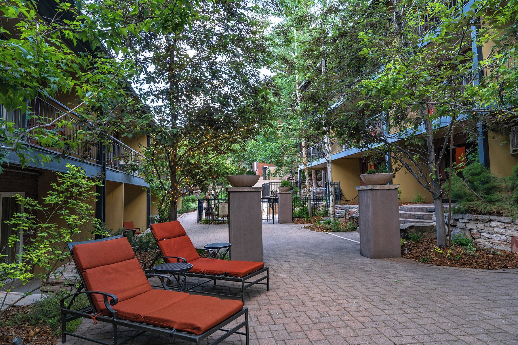 An outdoor seating area with lounge chairs and tables, surrounded by trees and a stone pathway.