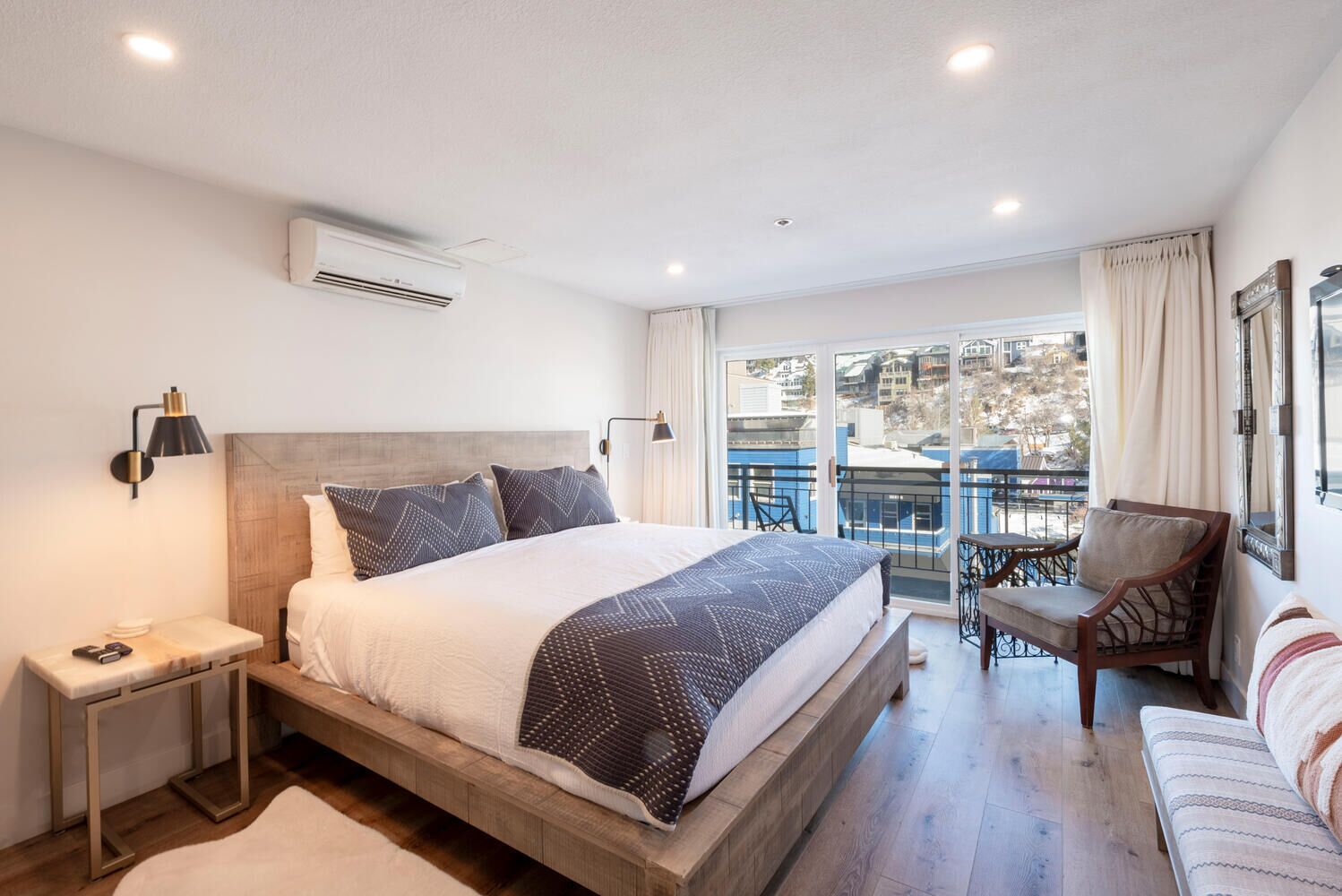 A bedroom with a large bed, a nightstand, a chair, a sofa, and a balcony with a view of buildings.