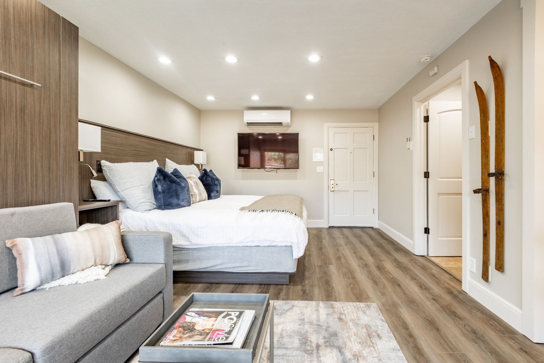 Quarto moderno com cama, sofá cinzento, mesa de centro com revistas, televisão e ar condicionado.
