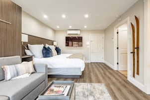 Quarto moderno com cama, sofá cinzento, mesa de centro com revistas, televisão e ar condicionado.