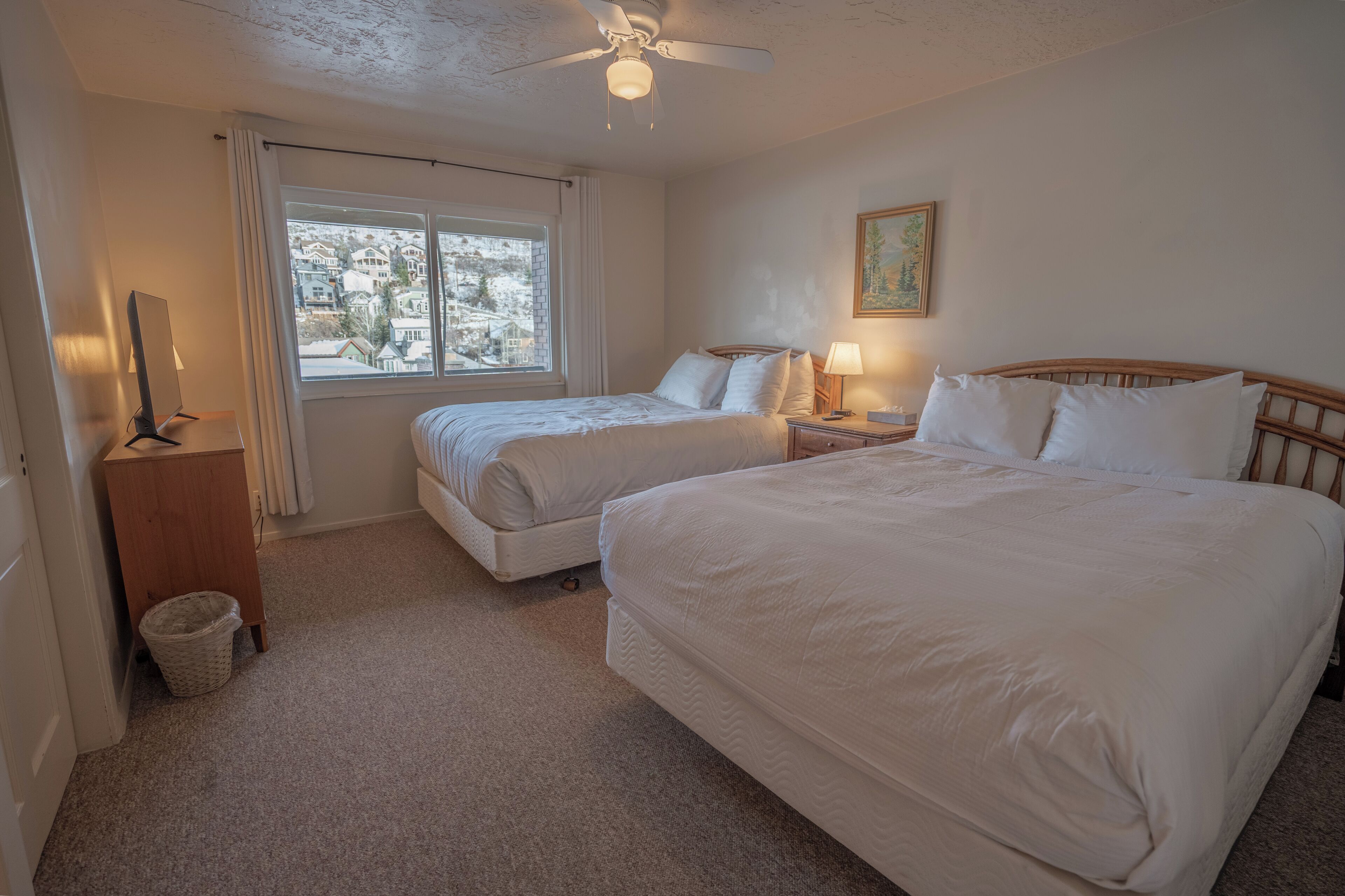 A hotel room with two beds, a ceiling fan, a window with a view of houses, a lamp, and a painting on the wall.