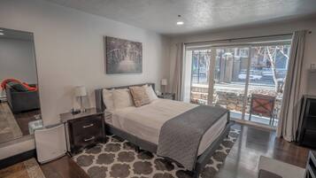 A modern bedroom with a large bed, bedside tables, a nightstand, a mirror, and a view of the outdoors through a sliding glass door.