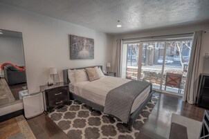 A modern bedroom with a large bed, bedside tables, a nightstand, a mirror, and a view of the outdoors through a sliding glass door.