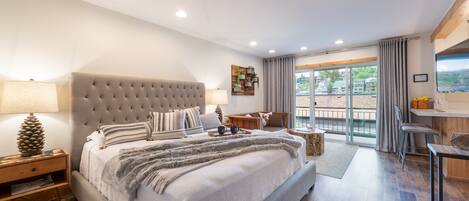 A modern bedroom with a large bed, a nightstand, a desk, and a view of the outdoors.