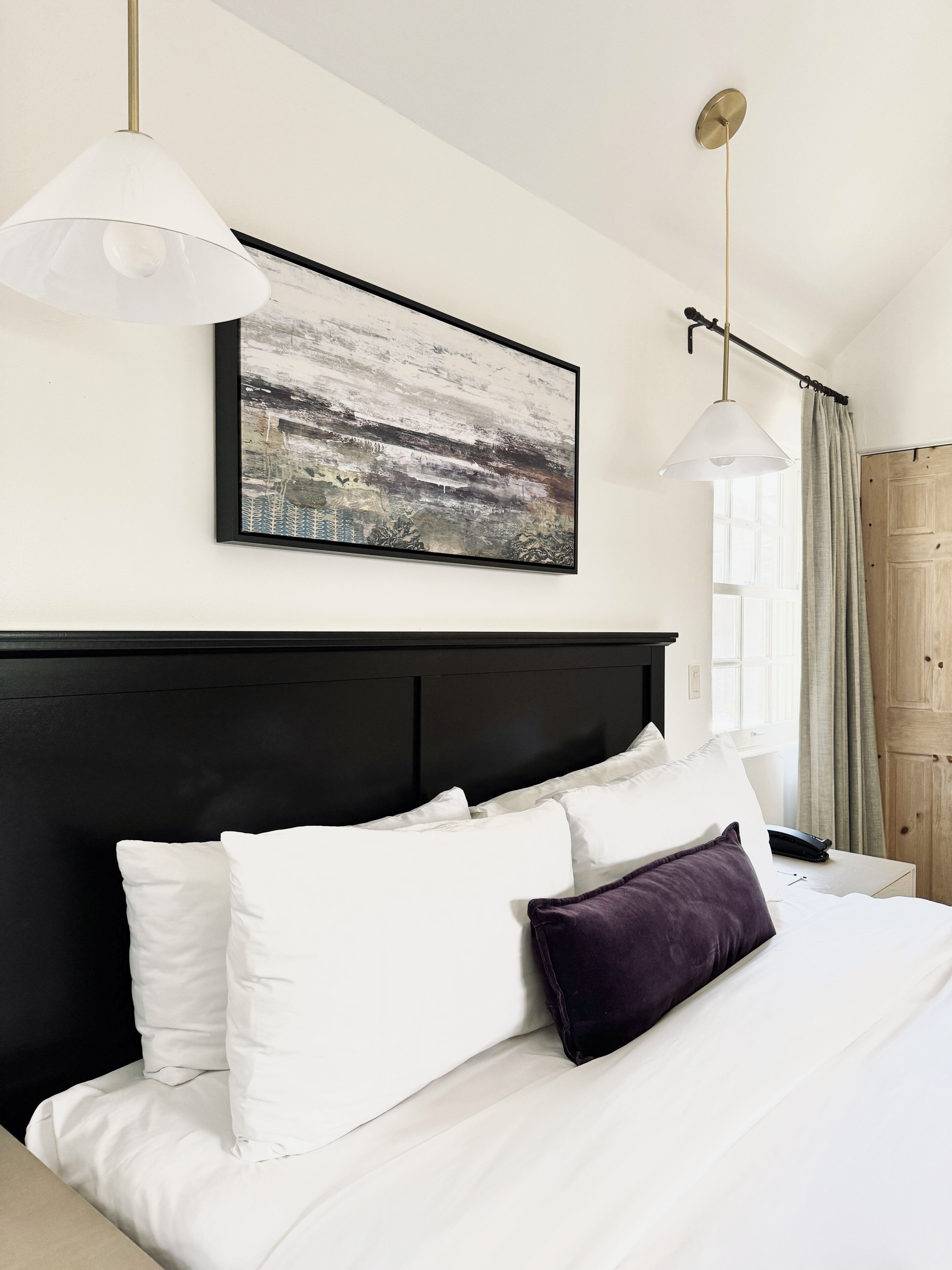 A hotel room with a large bed, white bedding, a black headboard, a framed abstract painting, and two pendant lights.