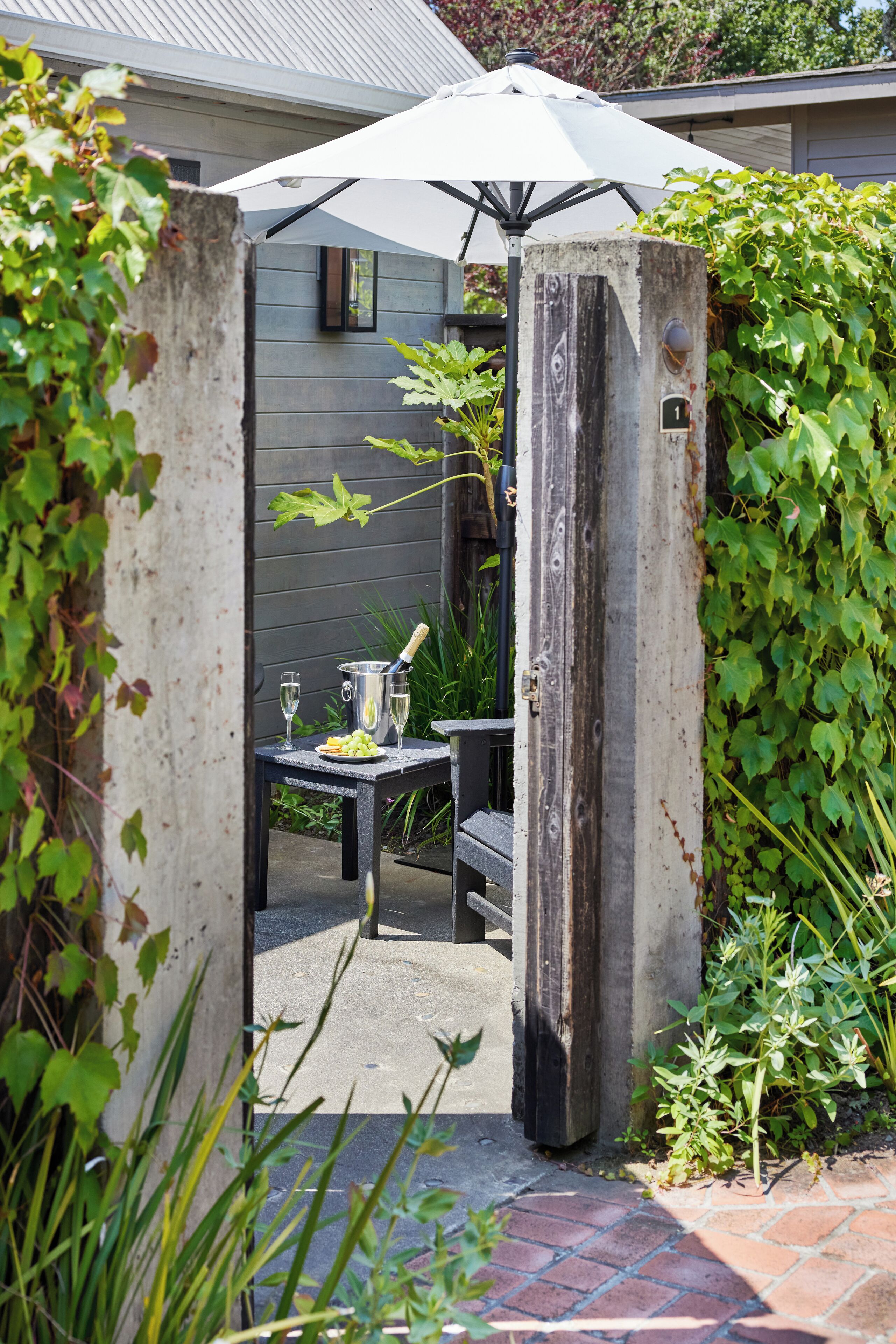 Un coin patio aménagé dans le jardin, avec une table pour deux personnes, un parasol blanc et un poteau en bois portant un numéro.