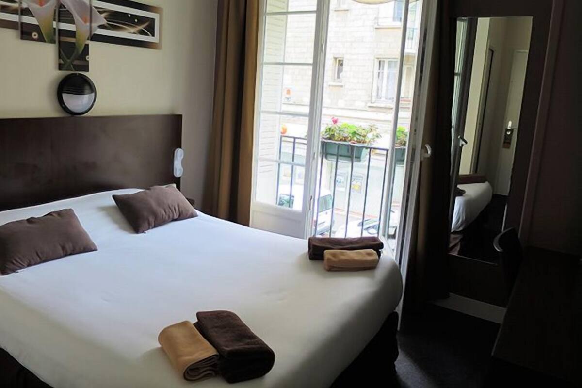 A hotel room with a large bed, two pillows, a bedside table, and a window with a view of the street.
