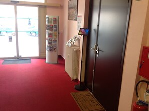 A hotel lobby with a red carpet, a bulletin board with various notices, a digital display, and a fire extinguisher. - Hotel de la Paix (Caen)