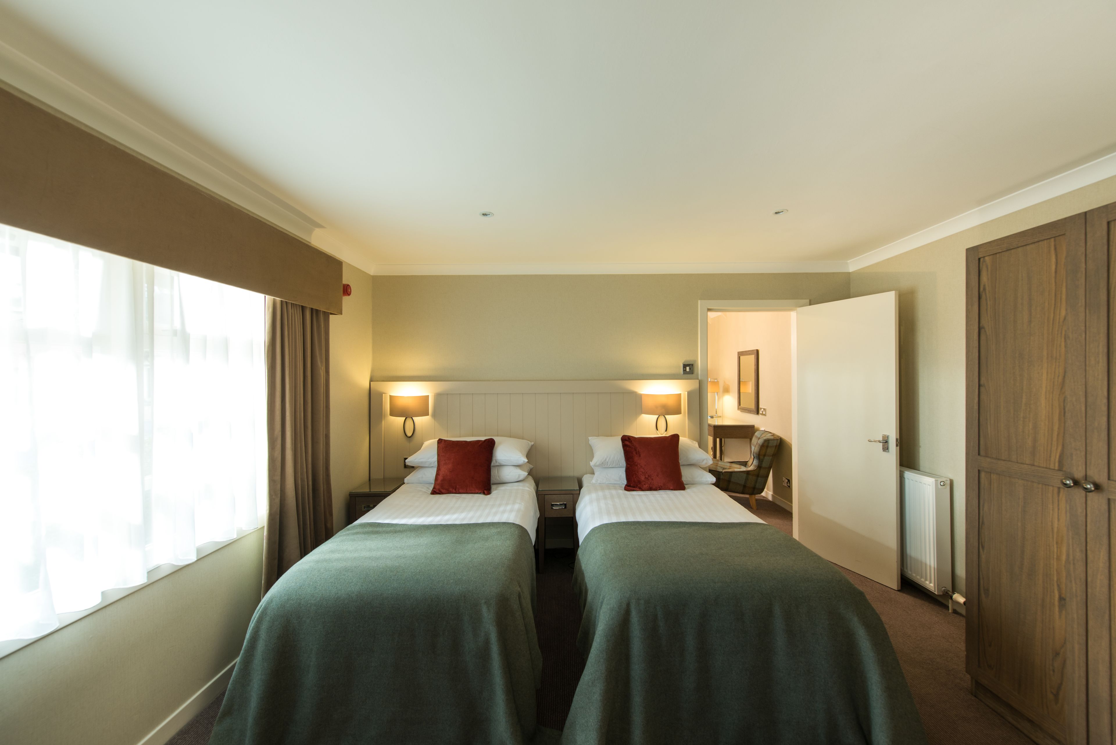 Habitación de hotel con dos camas individuales, una mesita de noche, una lámpara, un armario y una ventana con cortinas.