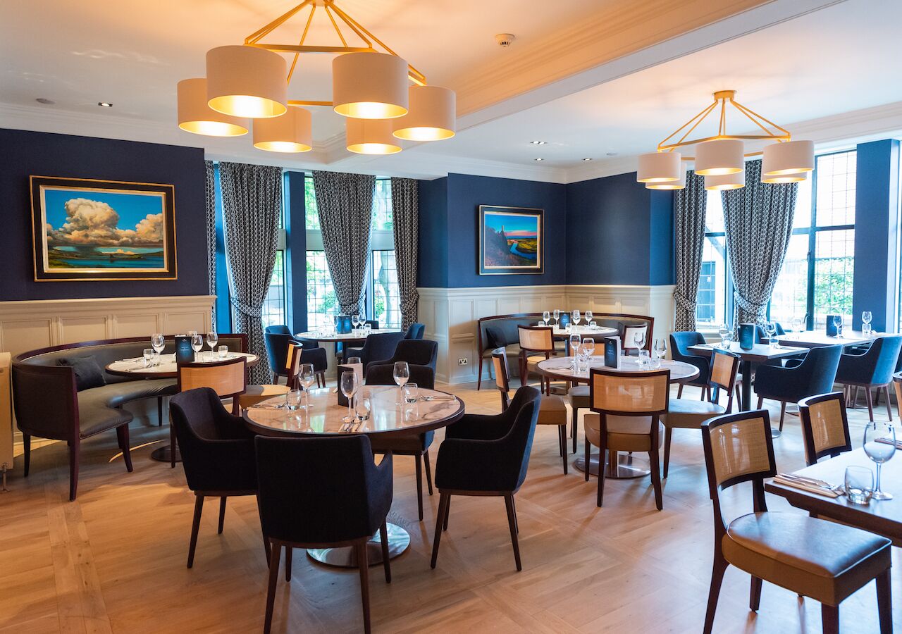 A restaurant with round tables and chairs, large windows with curtains, and artwork on the walls.