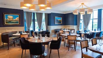 A restaurant with round tables and chairs, large windows with curtains, and artwork on the walls.