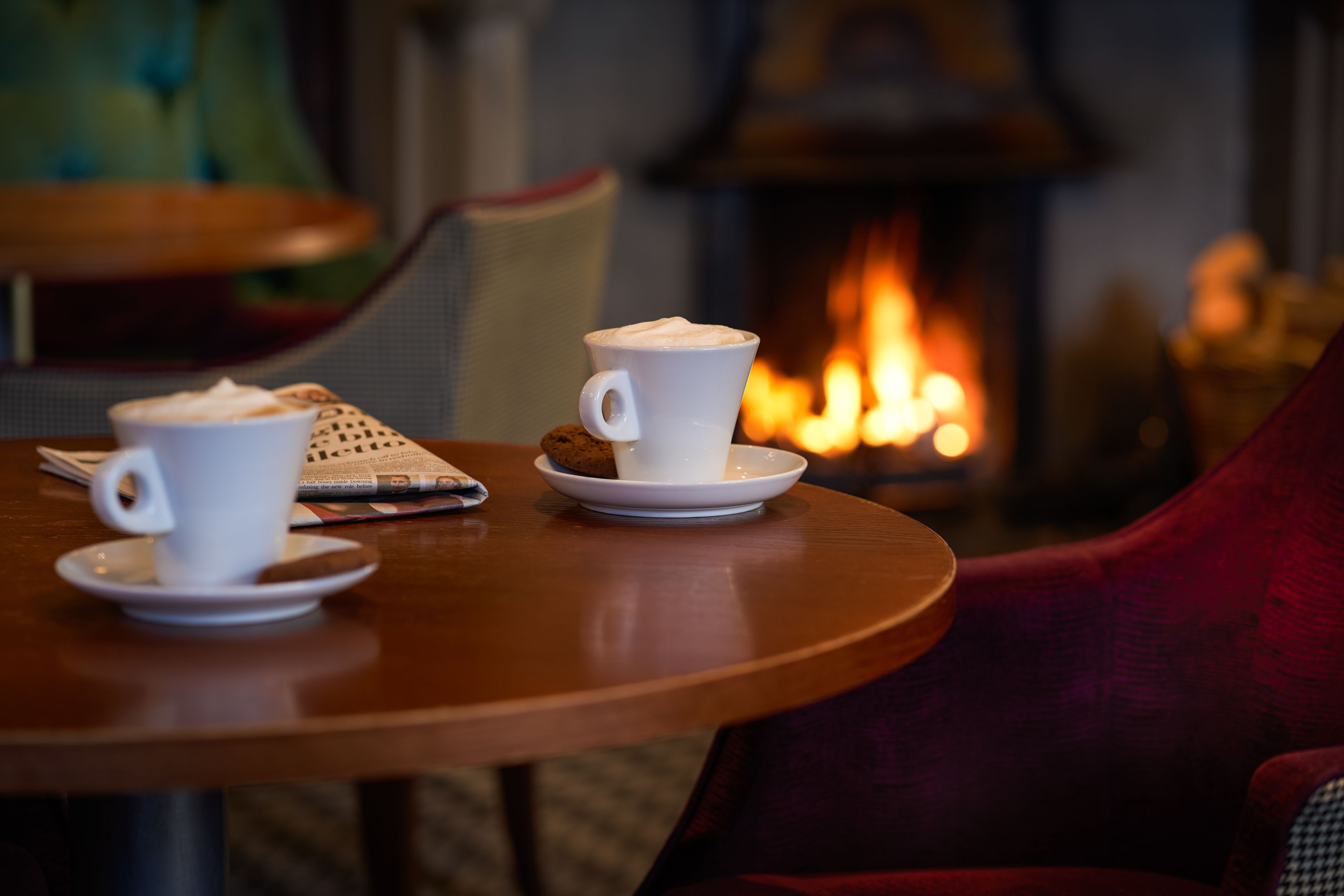 Un rincón acogedor con dos tazas blancas de café sobre platillos, un periódico y una chimenea al fondo.