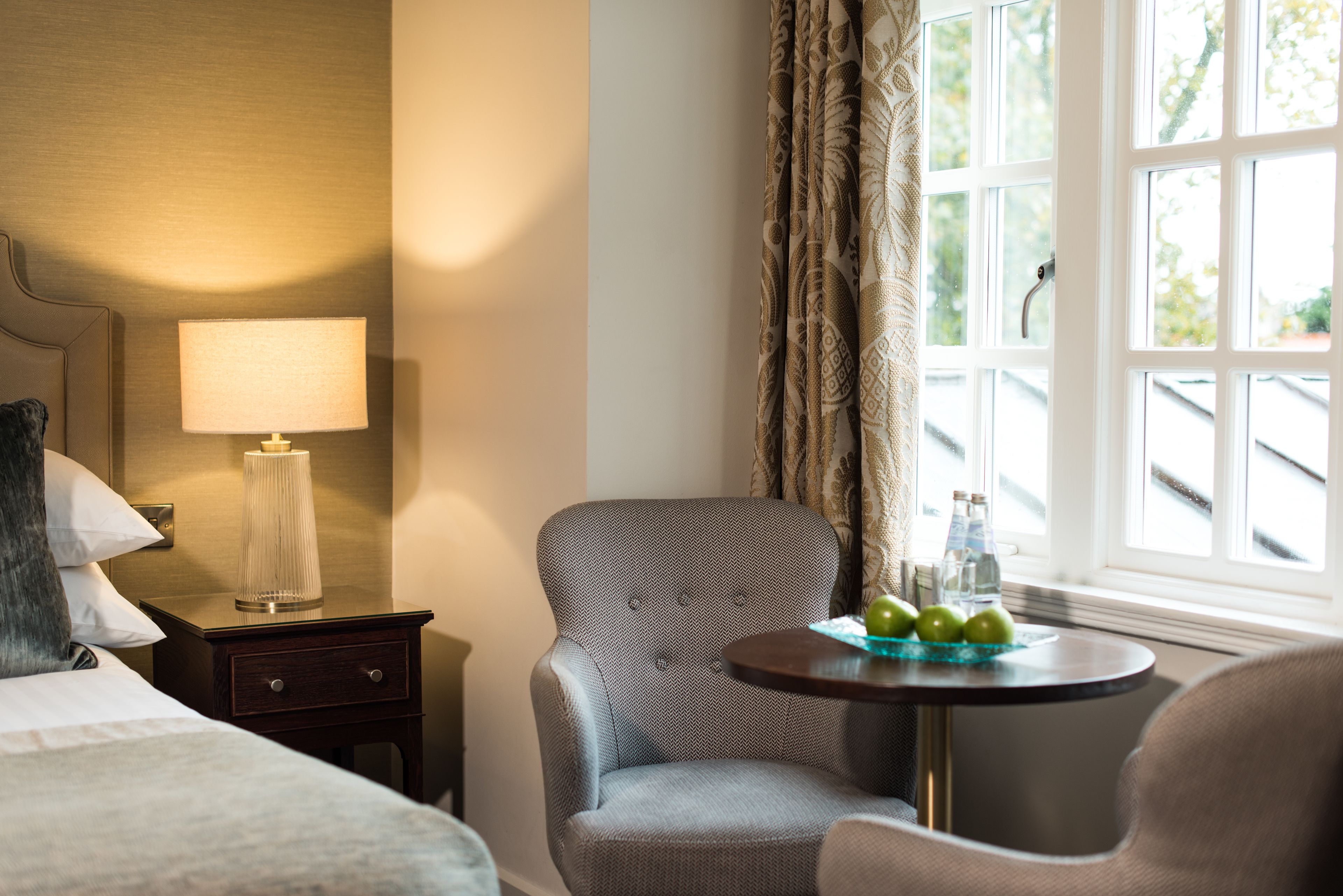Une chambre d’hôtel avec un lit, une table de chevet avec une lampe, une chaise, une petite table avec des pommes et une fenêtre avec des rideaux.