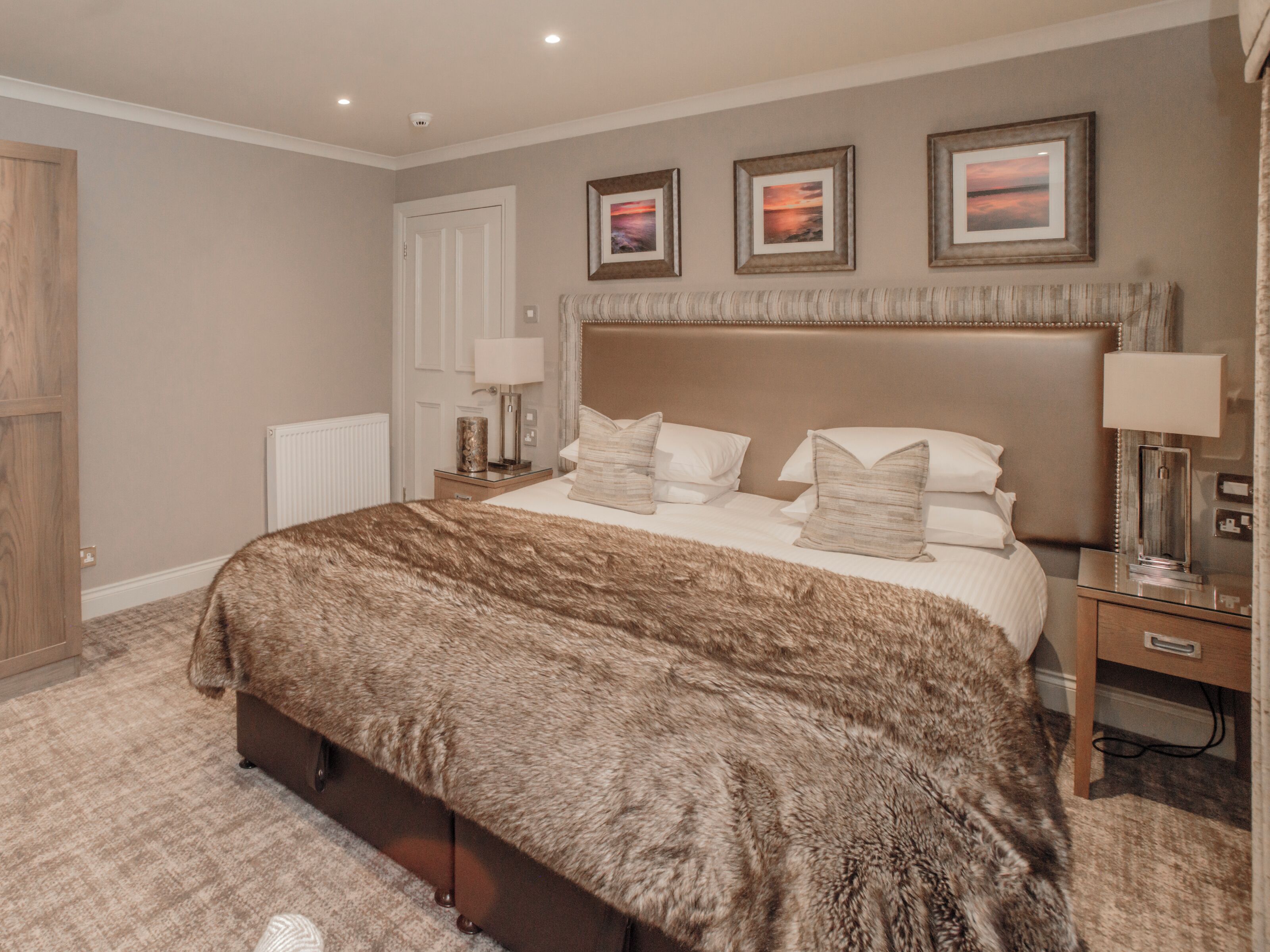 A bedroom with a large bed, a wooden wardrobe, a nightstand with a lamp, and three framed pictures on the wall.