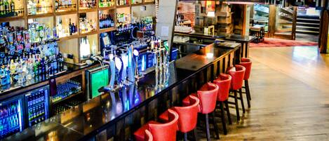 Zona de bar con una barra alargada, taburetes rojos y estantes llenos de diferentes botellas.
