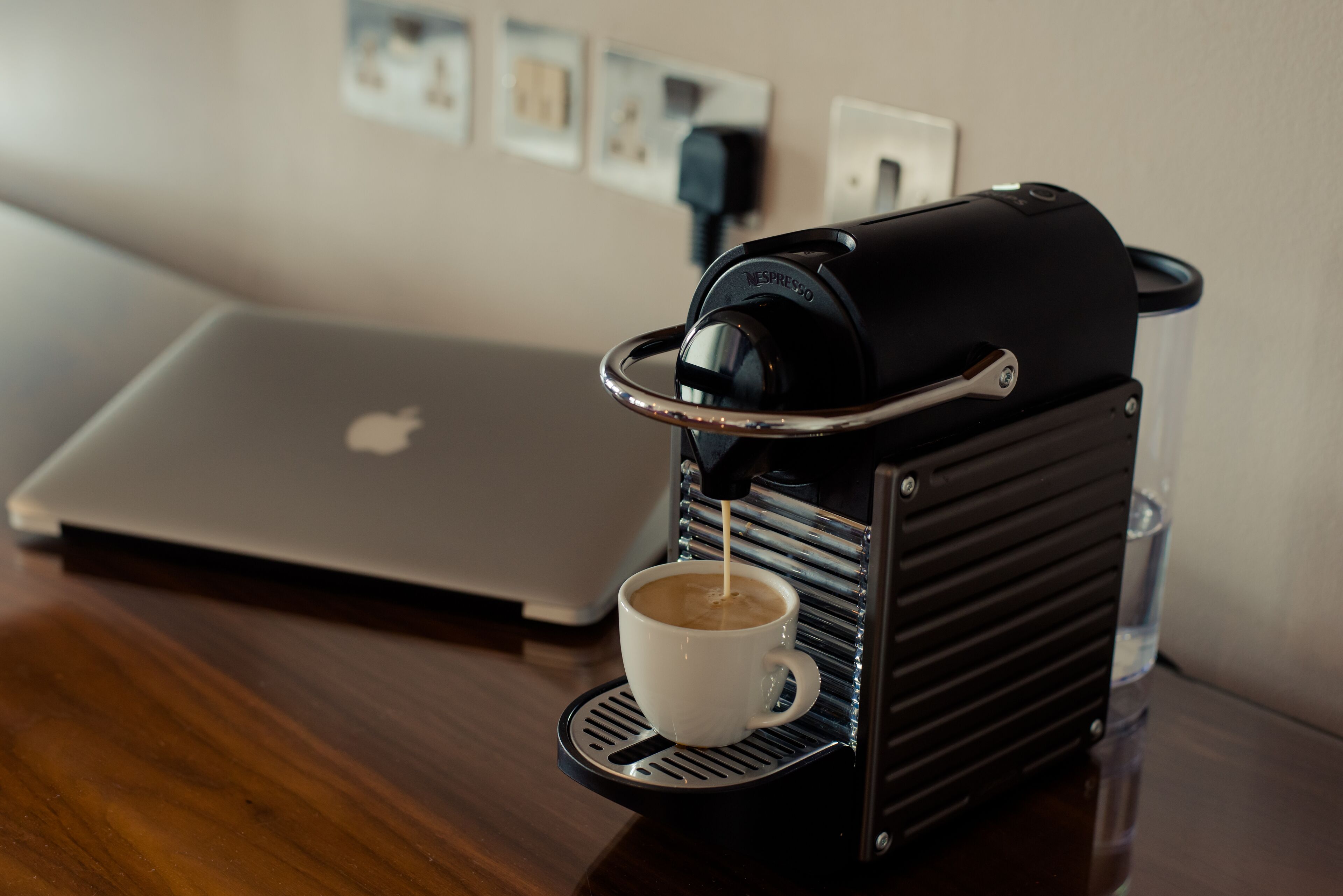 Een koffieapparaat schenkt koffie in een kop, een laptop en een waterdispenser staan op een houten tafel.