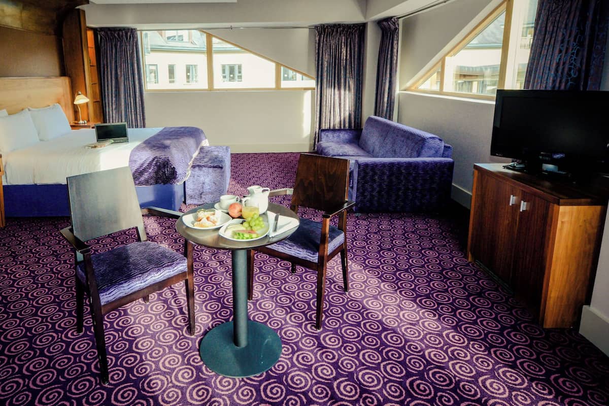 Habitación de hotel con cama, escritorio con computadora, televisor, dos sillas, una mesita con desayuno y alfombra estampada.