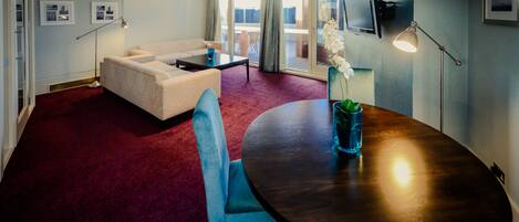 Habitación de hotel con alfombra roja, una mesa redonda de madera, un sofá blanco y un sillón azul. Hay un televisor montado en la pared y una ventana con cortinas.