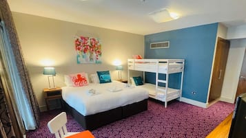 Habitación de hotel con literas, un escritorio, una silla, una cama con almohadas, un cuadro en la pared y una alfombra estampada.
