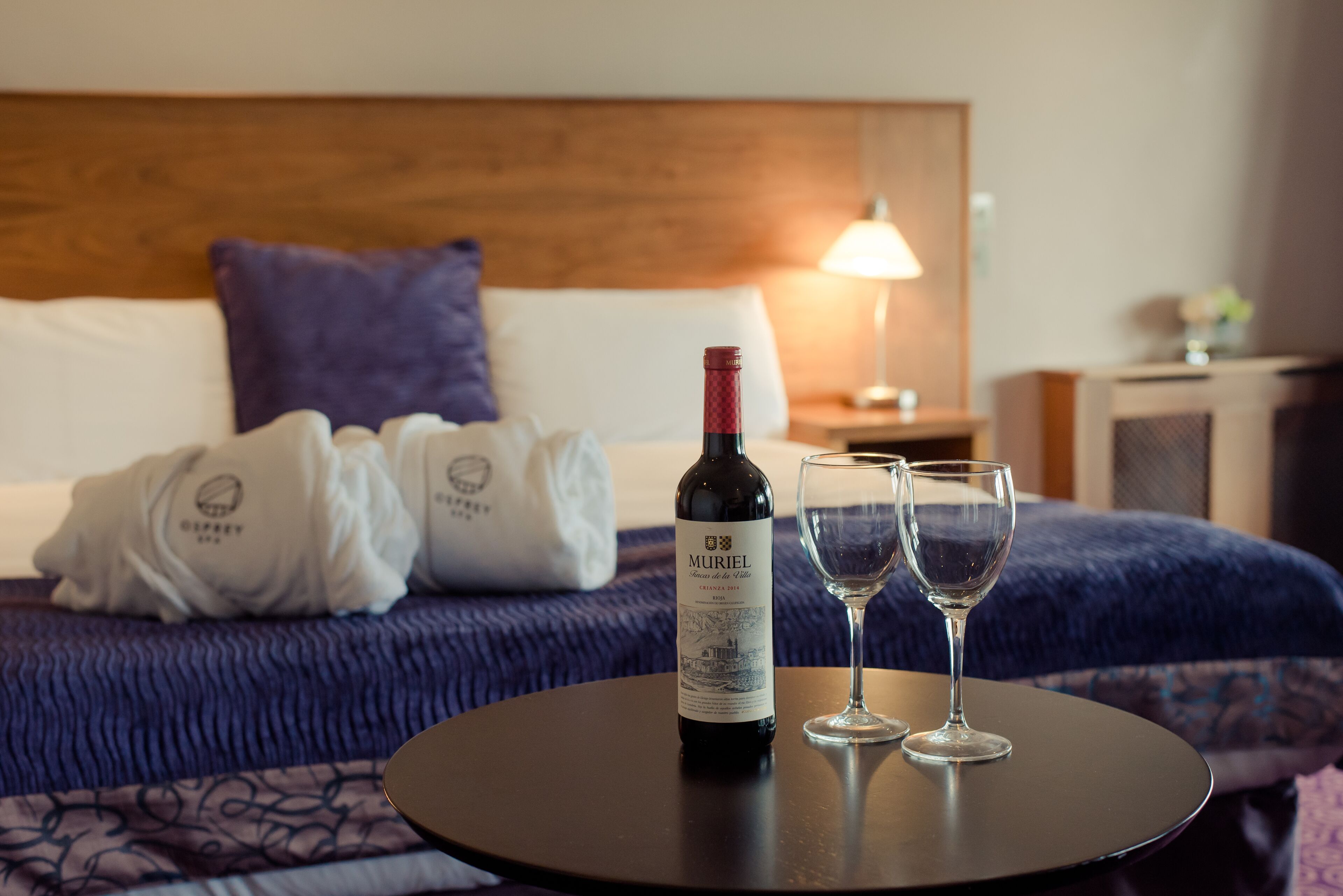 Quarto de hotel com uma cama, uma mesa redonda com uma garrafa de vinho e duas taças, e uma mesinha de cabeceira com um abajur.