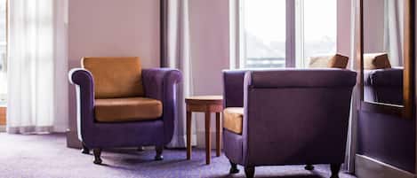A room with a purple carpet, two armchairs, a small wooden table, and a view through large windows.