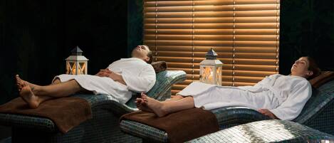 A spa room with reclining chairs, a window with blinds, and two lit lanterns.