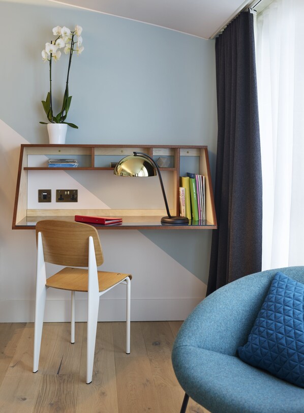 A modern workspace with a wooden desk, a white chair, a blue armchair, a desk lamp, a potted orchid, and a bookshelf. - Marrable's Farringdon Hotel (London)