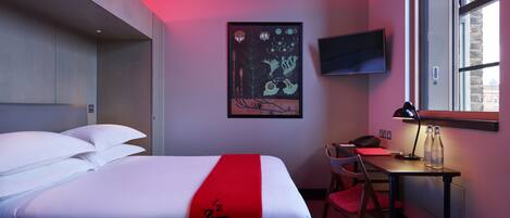 Una habitación de hotel con una cama, un escritorio con silla, un televisor en la pared y una ventana con vistas a edificios.