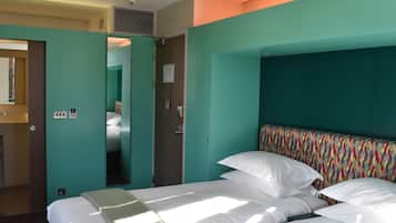 A hotel room with teal walls, a bed with white linens, a colorful headboard, and a mirror on the left wall.
